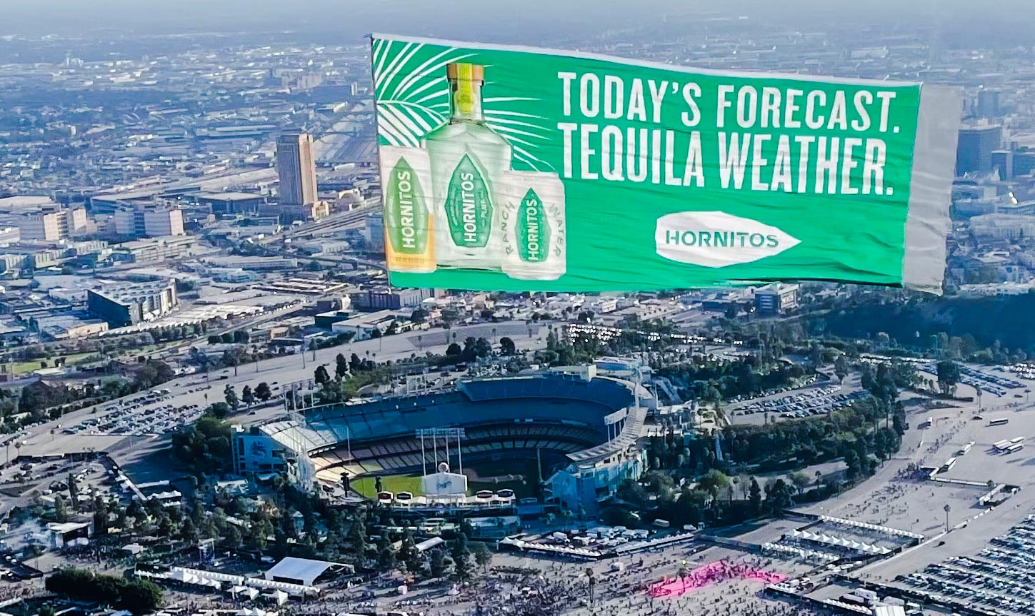 An overhead view of a bustling sports stadium and lively tailgate crowd, with a vibrant green aerial billboard featuring the Hornitos Tequila brand logo and a prominent image of a tequila bottle, adding excitement and energy to the atmosphere.
